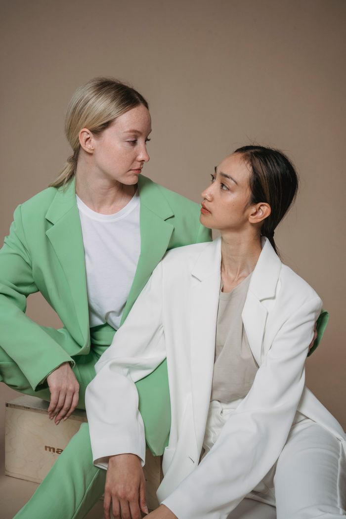 services-02 Two women in pastel blazers share a moment in a minimalist studio portrait.