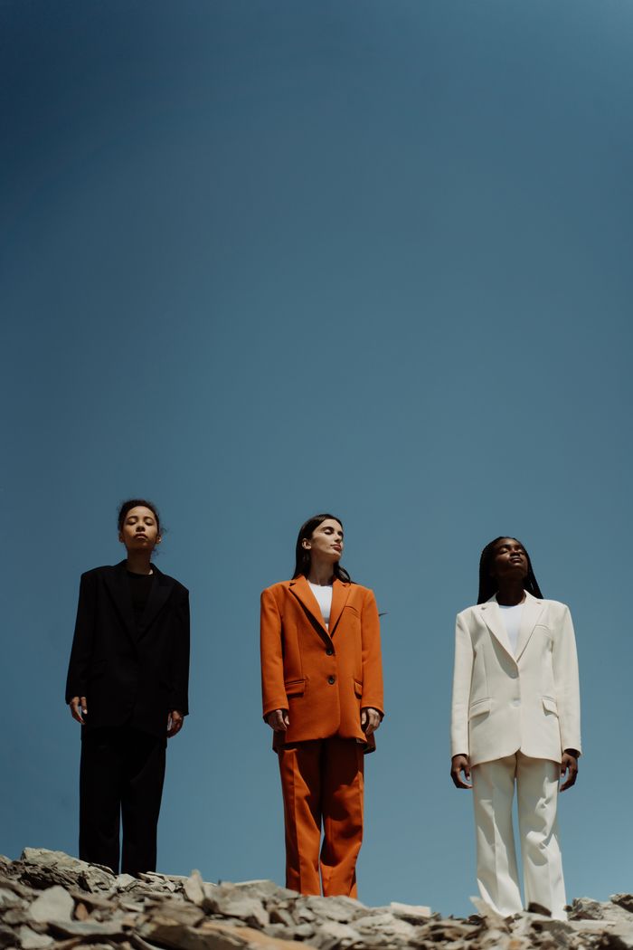 services-03 Three women wearing fashionable blazers stand confidently outdoors against a clear blue sky.